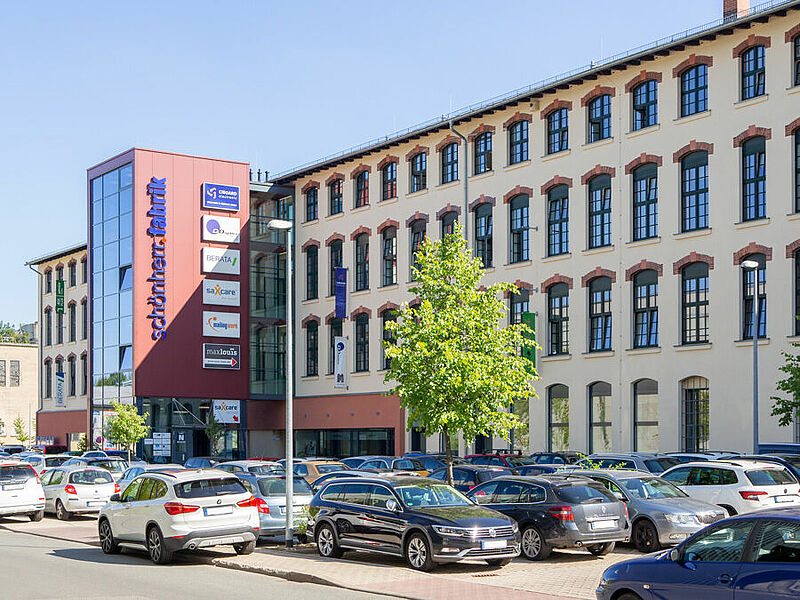 Das Bild zeigt ein Fabrikgebäude mit einem roten Anbau. Darauf steht Schönherrfabrik. Vor dem Haus ist ein großer Parkplatz mit vielen Autos. In der Mitte steht ein kleiner Baum.
