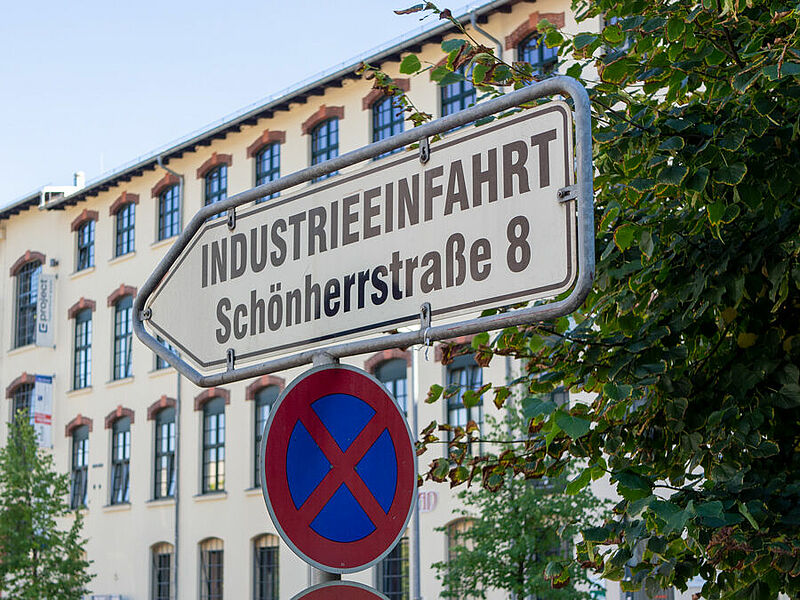 Das Bild zeigt ein Straßenschild mit der Aufschrift "Industrieeinfahrt Schönherrstraße 8". Im Hintergrund sieht man ein großes Fabrikgebäude. Unter dem Schild steht ein Halteverbotsschild und rechts ragt ein Busch ins Bild.